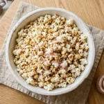 Overhead view of stovetop popcorn in a white bowl topped with pink Himalayan salt crystals and brown butter on a wooden surface